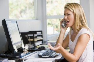 Woman in home office with computer using phone frowning
